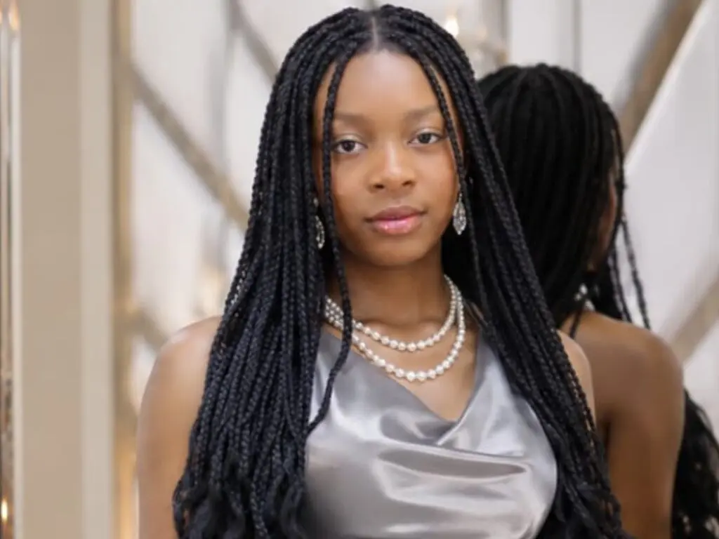 NaomiBennetti poses in a silver satin top with long black braids and pearl necklaces, looking directly at the camera.