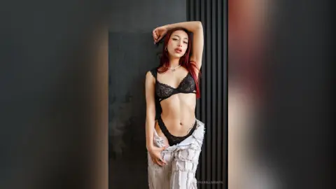 AuroraBellmontk poses in a black lace bra and matching thong, wearing white ripped jeans while showing her flat stomach and midriff against a dark wall.