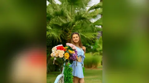Smiling woman in a blue summer outfit holding a colorful flower bouquet next to a bicycle in a lush garden.