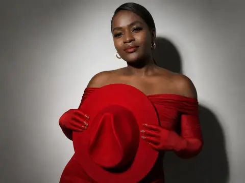 Elegant Black woman in a red off-the-shoulder dress holding a wide-brimmed red hat against a neutral background.