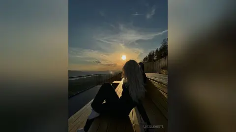 Blonde woman in black outfit sitting on a wooden deck overlooking a mountain valley at sunset.