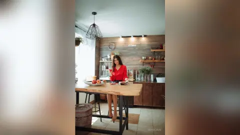 Elegant woman in a red blouse sitting at a wooden kitchen island with fruit in a cozy, modern home setting.