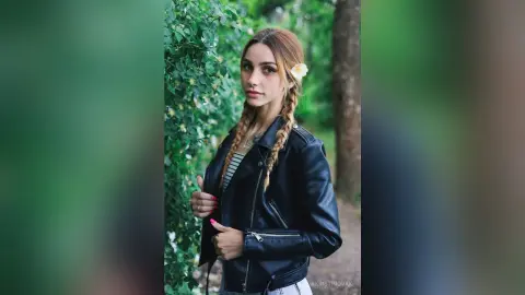 Young woman in a black leather jacket with braided hair posing in a lush green forest setting.