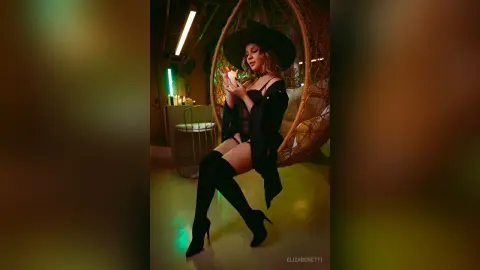 Stylish woman in a black hat and chic outfit sitting in a hanging egg chair within a moody, neon-lit room.