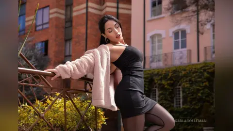 Elegant woman in a black dress and fuzzy pink cardigan posing on a city balcony during golden hour.
