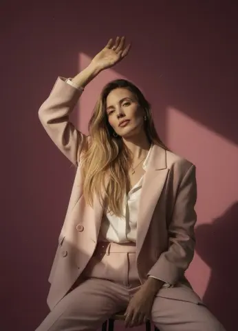 Elegant woman in a blush pink suit posing against a pink background with dramatic sunlight shadows.