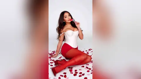 Glamorous woman in a white corset and red skirt sitting against a white background with a playful mood.