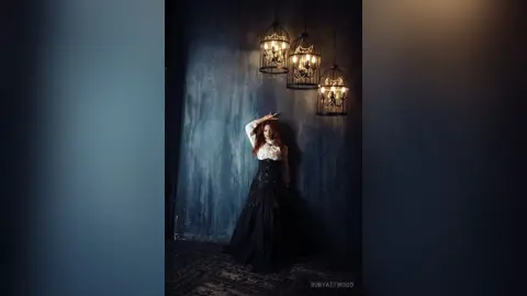 RubyAttwood stands against a dark blue wall wearing a black corset and a long, full black skirt.