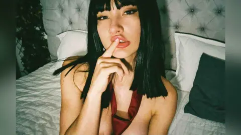 Young woman with dark hair in a red top posing on a white bed in a moody, intimate bedroom setting.