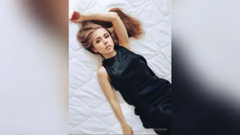 Elegant woman in a black satin dress lying on a white quilted bed in a serene, minimalist setting.