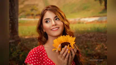 ValeryRouge smiles while holding a bright yellow sunflower near her face against a soft, natural outdoor background.