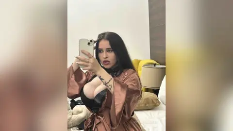 Young woman in a brown satin robe taking a selfie in a cozy bedroom setting.