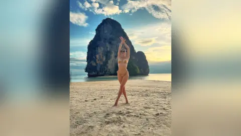 AnaMathers stands on a sandy beach wearing a tan bikini with her arms raised above her head against a large rock formation.