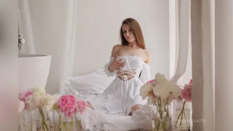 IngridBeckton poses in a white lace top and skirt while sitting on a bed surrounded by pink and white flowers.