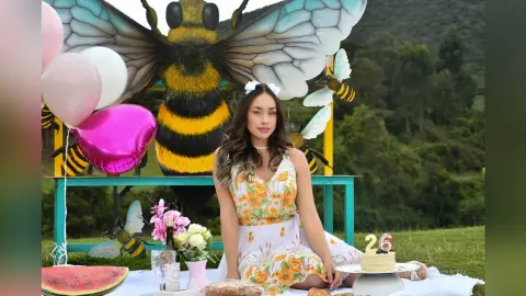 Young woman in a floral sundress celebrating a 26th birthday picnic with giant bee decorations in a green field.