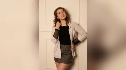 Young woman in a stylish black top, white blazer, and plaid skirt posing against a white door with a soft mood.