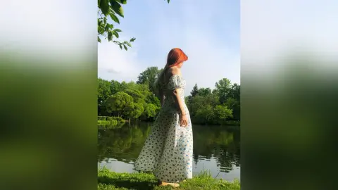 Redheaded woman in a white floral maxi dress standing by a peaceful lake in a lush green forest.