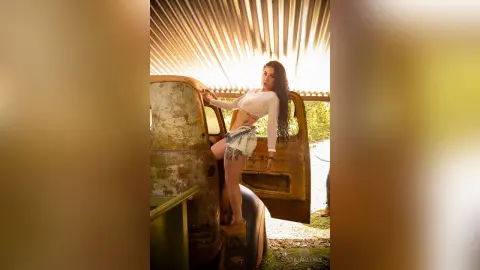 JuliahLewis leans against a rusted vintage truck wearing a white long-sleeve crop top and denim cut-off shorts.