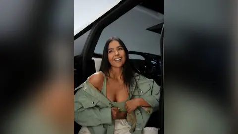 Smiling young woman in a sage green jacket sitting in a car during a bright daytime drive.