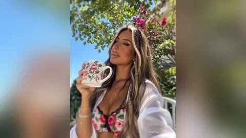 Young woman in a white robe holding a floral teacup outdoors near blooming pink flowers on a sunny day.