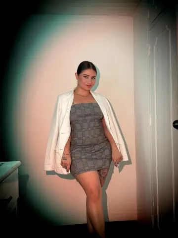 Smiling woman in a grey plaid mini dress and white blazer posing in a soft-lit indoor setting.