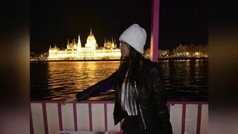 LucyMartin poses in a black leather jacket and white knit beanie against a lit background of a river and a grand building at night.