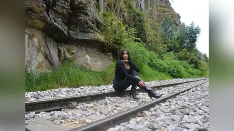 AmeliaStell sits on a gravel railway track wearing a black long-sleeved top and black leggings with black boots against a rocky cliffside.