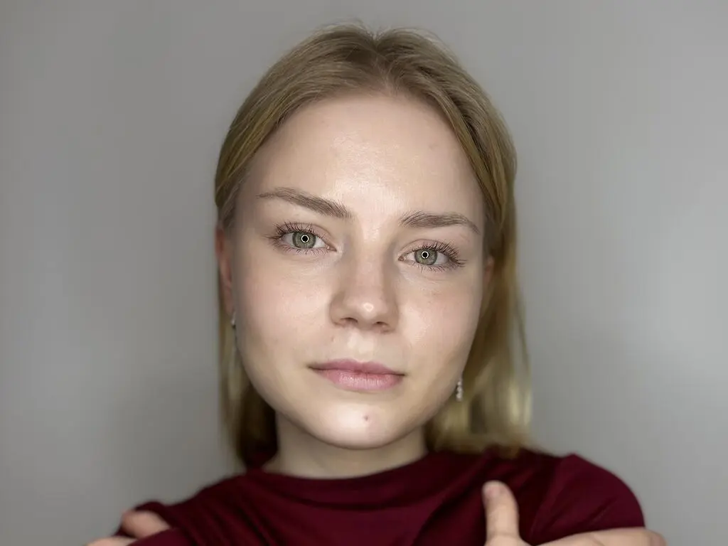 Close-up portrait of a blonde woman with natural makeup wearing a maroon top against a neutral background.