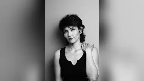 IveteDurand poses looking at the camera, wearing a black tank top with her hand resting on her shoulder against a plain background.