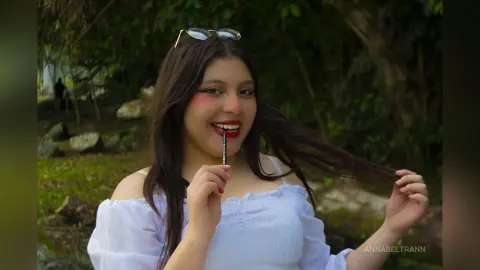 Smiling young woman in a white off-the-shoulder top applying lip gloss in a lush green outdoor garden setting.