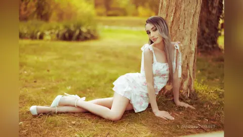MaddyScarlett sits on the grass next to a tree trunk wearing a white floral sundress and white high heels.