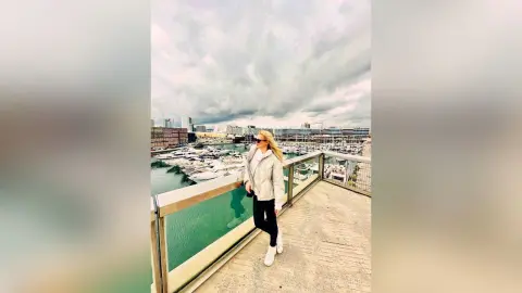 MilenaBia stands on a balcony overlooking a marina, wearing a white jacket and black leggings.