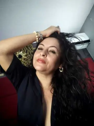 Smiling woman with long dark hair wearing a black top and gold jewelry in a cozy indoor setting.