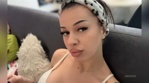AriaVex wears a white patterned headband and a white lace bra while sitting on a dark grey sofa.