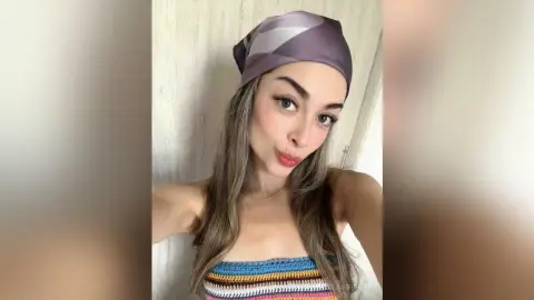 Young woman with long hair wearing a purple headscarf and colorful striped tube top in a bright indoor setting.