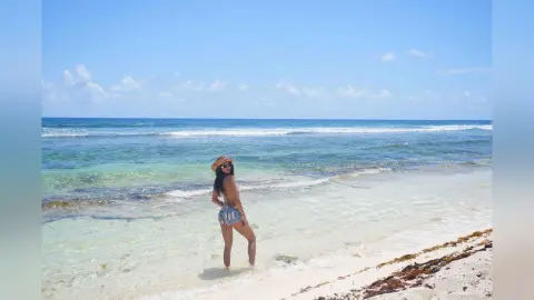 LucyMartin stands in the shallow ocean water wearing a blue and white patterned bikini and a straw sun hat.
