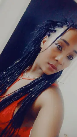 Young woman with long black braids wearing an orange top in a moody indoor setting.