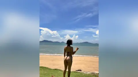 Woman in black bikini standing on a sunny beach looking out at the ocean and distant mountains.