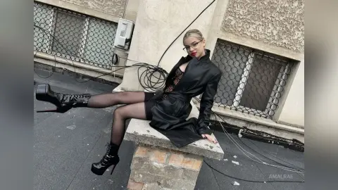 Stylish woman in a black leather jacket and sheer tights sitting on a ledge against a textured building wall.