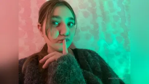 MartineKibbey poses with a finger to her lips, wearing a dark fuzzy sweater against a pink and teal lit background.