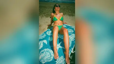 Smiling woman in a green bikini relaxing on a blue patterned beach towel under bright summer sunlight.