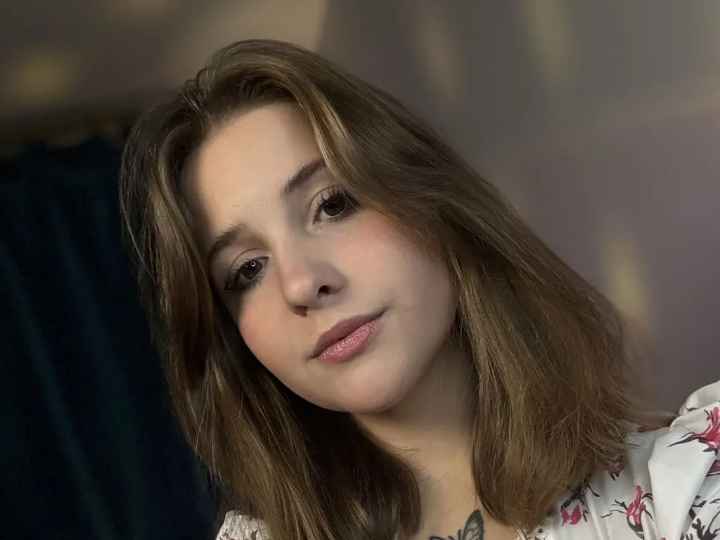 Young woman with long brown hair wearing a floral blouse in a soft, moody indoor setting.
