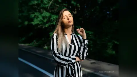 Serene woman in a black and white striped shirt standing on a road against a lush green forest background.