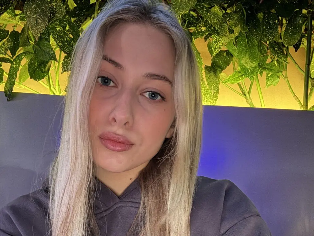 Blonde woman in a grey sweatshirt posing against a lush green leaf background with warm ambient lighting.