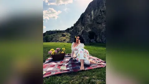 Woman in white floral dress sitting on a picnic blanket with flowers near a large mountain under a blue sky.