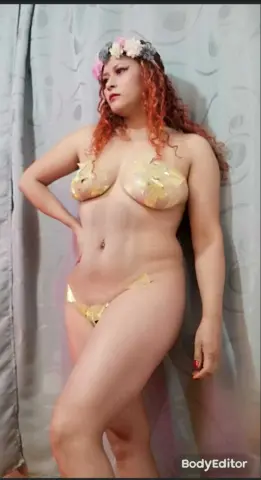 Redheaded woman in gold leaf bikini and floral crown posing against a grey curtain in soft lighting.