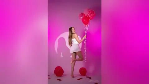 ArinnaGreen poses in a short white dress, holding red balloons against a pink background while showing off her long brown hair and legs.
