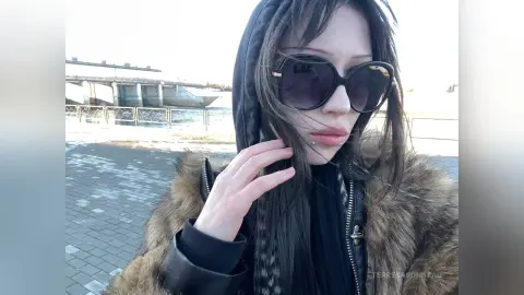Young woman with long hair wearing a black hoodie, fur coat, and sunglasses outdoors in a cool, moody setting.
