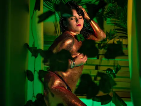 Woman with shimmering gold body glitter posing amidst lush green tropical leaves in moody emerald lighting.