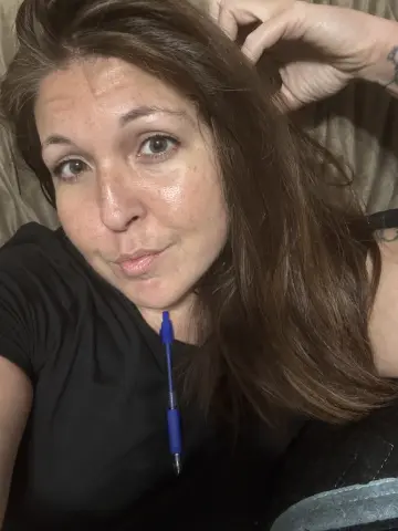 MistressBitchcoin looks at the camera with her long brown hair down, wearing a black t-shirt while sitting on a couch.